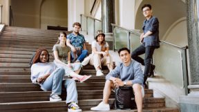Sechs internationale Studierende sitzen entspannt gemeinsam in der Hoschule auf einer Treppe und lächeln in die Kamera.